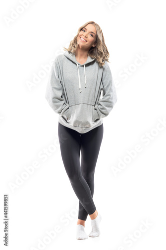 Happy relaxed young woman in hooded sweatshirt smiling and posing with hands in pockets. Full body length isolated on white background.