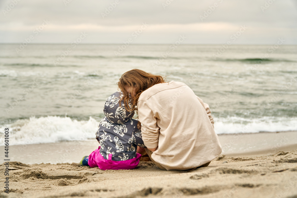 Mom and daughter are sitting together on the beach and hugging. Cold sea in autumn or winter. Breathe sea air by the ocean. Clouds and blue sky. View from the back. jumpsuit for a walk.