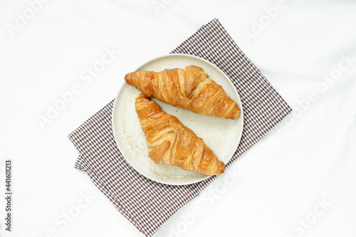 top view of  Two croissants are placed in a round ceramic  plate placed on brown checkered fabric against white crumpled tablecloths with soft light in the morning.