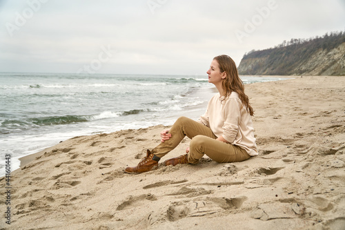 A beautiful woman sits on the sand on the beach and looks into the distance of the sea at the ocean horizon. Fall clothes vacation. Caucasian girl is happy Beige clothes and sand in the fall.