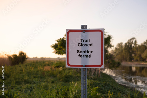 Sign trail closed in English and French at the park entrance at sunset time. Closure of outdoor facilities, national forests and provincial parks during novel corona virus outbreak quarantine.
