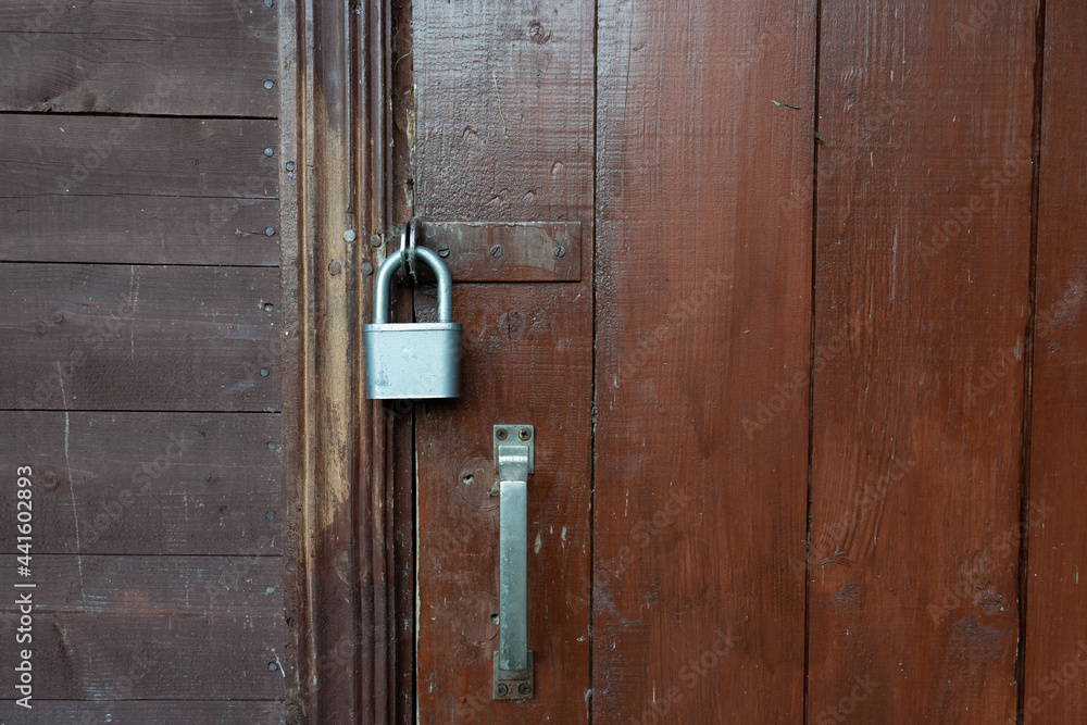 The brown wooden door of the village house is padlocked. Part of the wall is visible, sheathed with horizontal boards. Background. Texture. The brown wooden door of the village house is padlocked. Part of the wall is visible, sheathed with horizontal boards. Background. Texture.