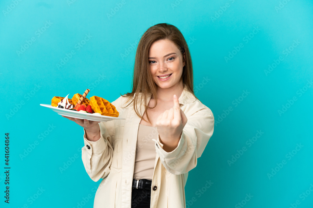 Teenager girl holding waffles over isolated blue background doing coming gesture Teenager girl holding waffles over isolated blue background doing coming gesture