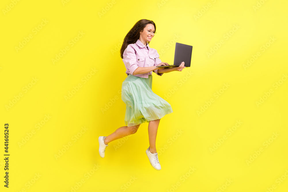 Full length body size photo portrait girl in skirt jumping high typing on laptop isolated bright yellow color background Full length body size photo portrait girl in skirt jumping high typing on laptop isolated bright yellow color background