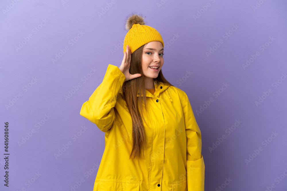 Teenager girl wearing a rainproof coat over isolated purple background listening to something by putting hand on the ear Teenager girl wearing a rainproof coat over isolated purple background listening to something by putting hand on the ear