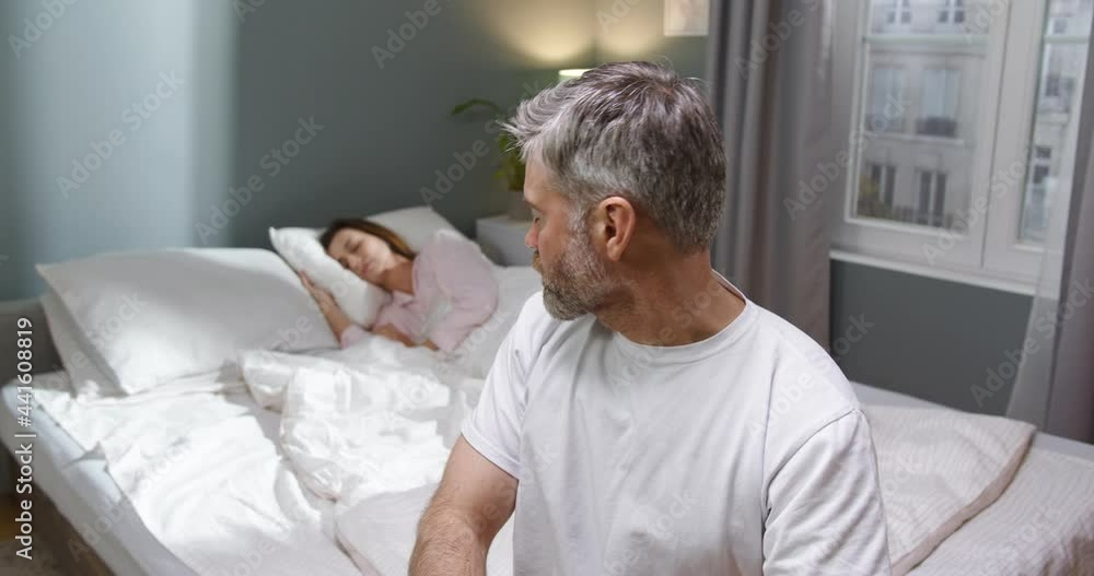 So upset. Waist up portrait view of the sad bearded man sitting at the bed and looking at his sleeping wife after the quarrel. Family disagreements concept So upset. Waist up portrait view of the sad bearded man sitting at the bed and looking at his sleeping wife after the quarrel. Family disagreements concept