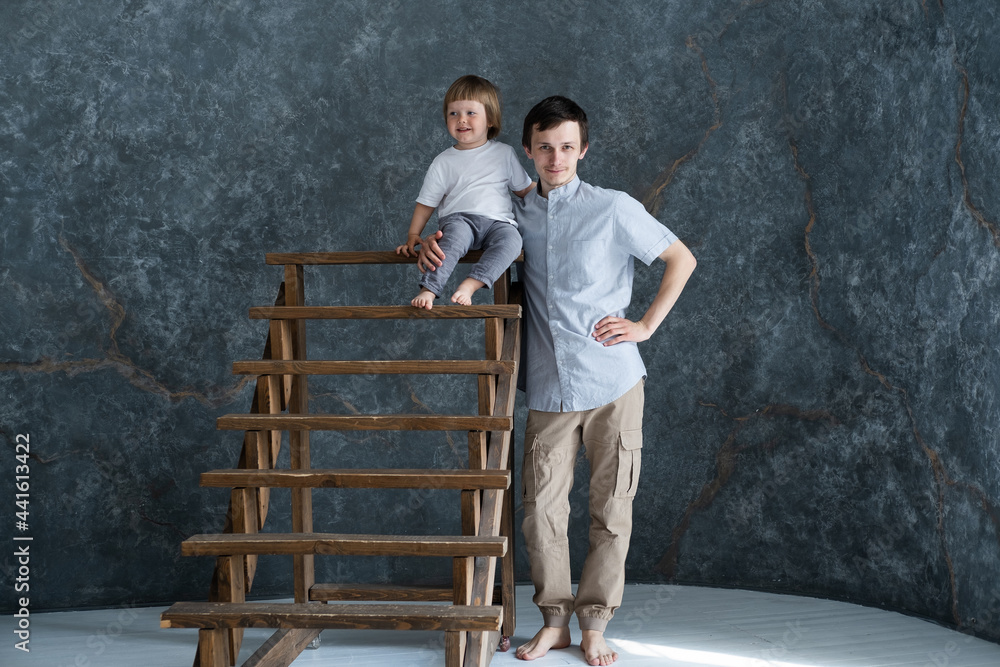 Studio portrait of Young father with his little son sitting on wooden stairs, grey background Studio portrait of Young father with his little son sitting on wooden stairs, grey background