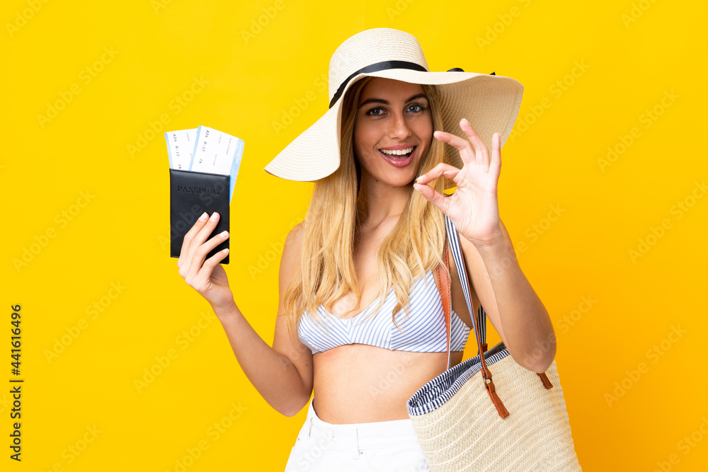 Young Uruguayan blonde woman in swimsuit holding a passport over isolated yellow background showing ok sign with fingers Young Uruguayan blonde woman in swimsuit holding a passport over isolated yellow background showing ok sign with fingers