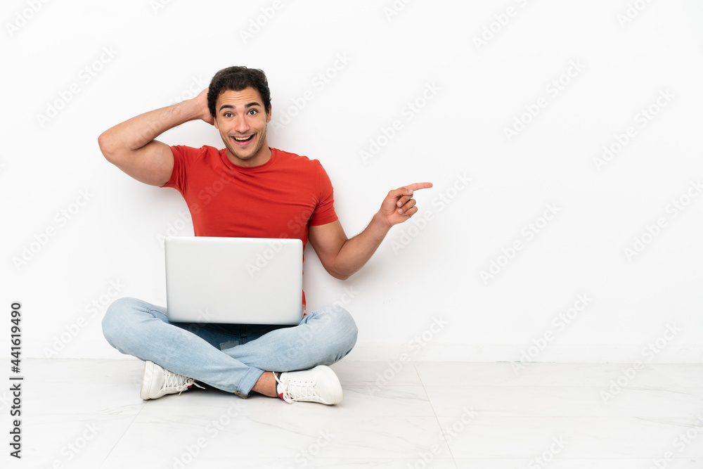 Caucasian handsome man with a laptop sitting on the floor surprised and pointing finger to the side Caucasian handsome man with a laptop sitting on the floor surprised and pointing finger to the side