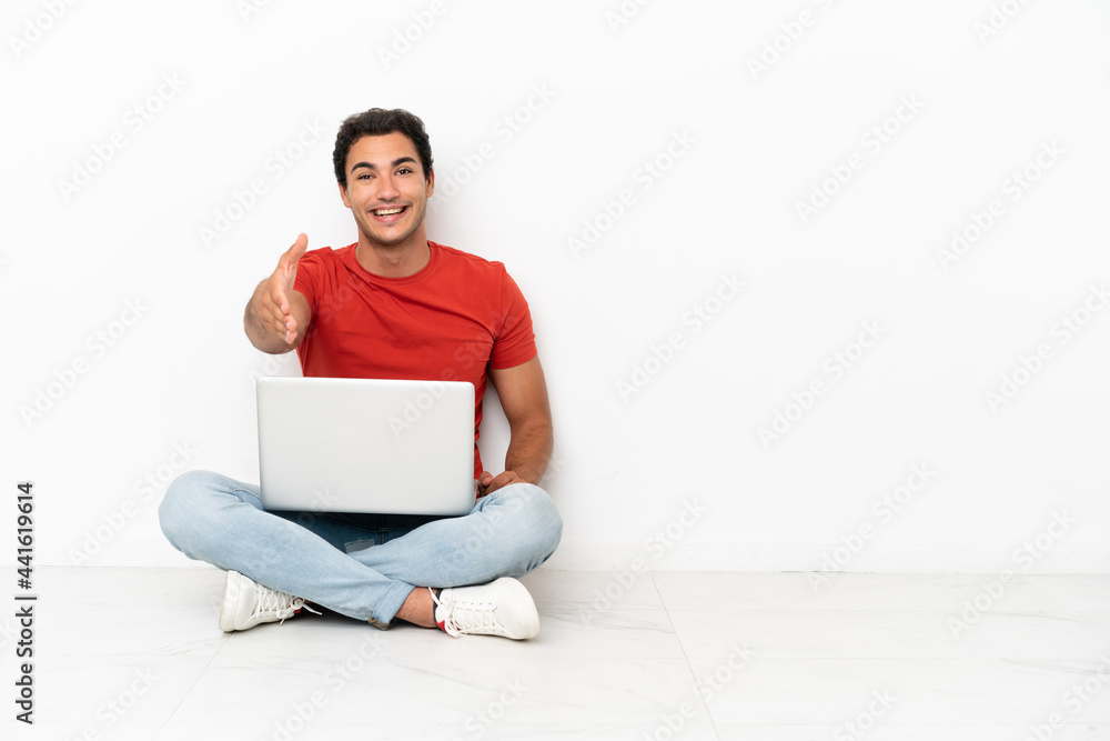 Caucasian handsome man with a laptop sitting on the floor shaking hands for closing a good deal Caucasian handsome man with a laptop sitting on the floor shaking hands for closing a good deal