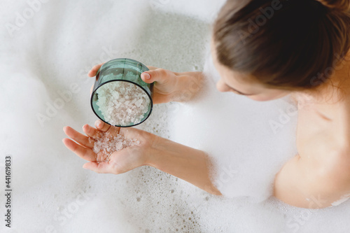 Top view of woman sitting in foam bath with sea bath salt in hands.