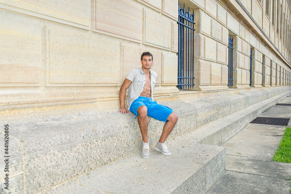 Dressing in a iron short sleeve shirt, blue shorts and sneakers , a young attractive guy is sitting against a pattern wall and relaxing.