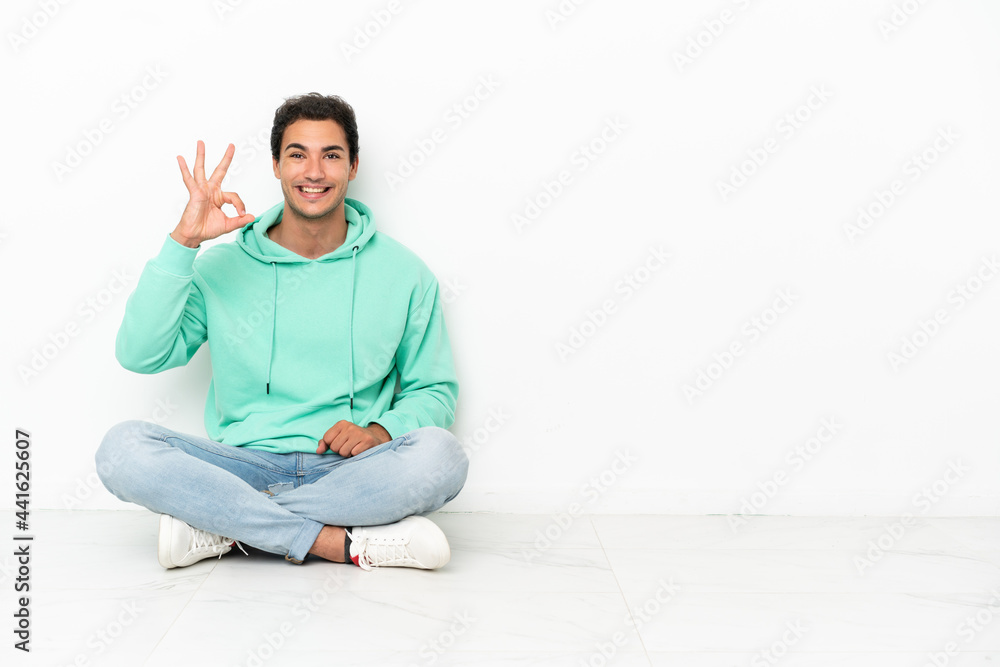 Caucasian handsome man sitting on the floor showing ok sign with fingers