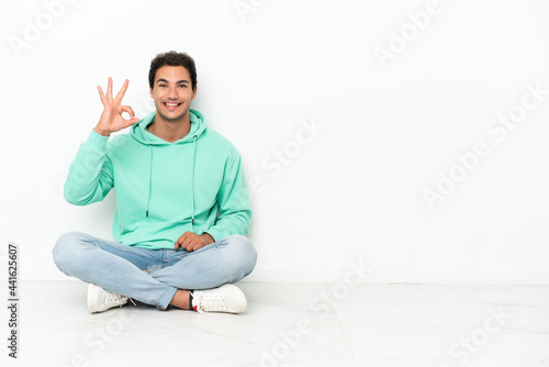 Caucasian handsome man sitting on the floor showing ok sign with fingers