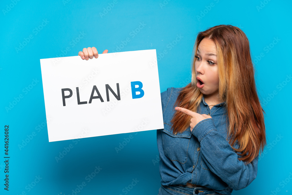 Teenager girl over isolated blue background holding a placard with the message PLAN B with surprised expression Teenager girl over isolated blue background holding a placard with the message PLAN B with surprised expression