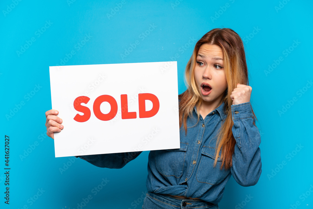 Teenager girl over isolated blue background holding a placard with text SOLD and celebrating a victory Teenager girl over isolated blue background holding a placard with text SOLD and celebrating a victory