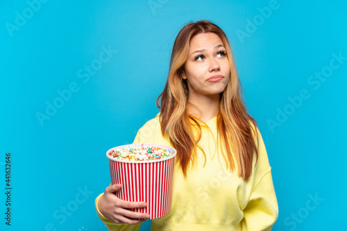 Teenager girl holding popcorns over isolated blue background and looking up