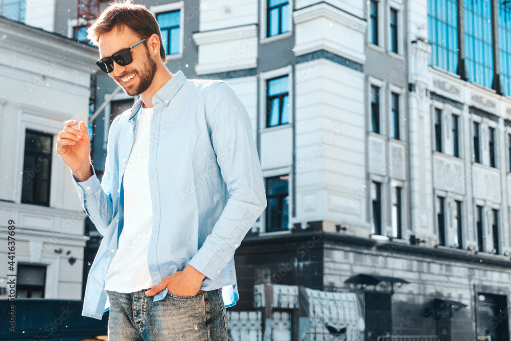 Portrait of handsome smiling stylish hipster lambersexual model.Modern man dressed in blue shirt. Fashion male posing near skyscraper on the street background in sunglasses. Outdoors at sunset Portrait of handsome smiling stylish hipster lambersexual model.Modern man dressed in blue shirt. Fashion male posing near skyscraper on the street background in sunglasses. Outdoors at sunset