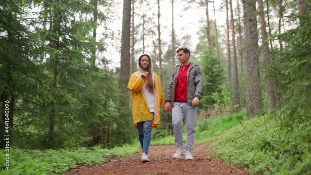 Low angle view of young couple of hikers walking down forest trail holding hands, woman holding and smelling flower. Happy romantic couple talking and having fun in travel Low angle view of young couple of hikers walking down forest trail holding hands, woman holding and smelling flower. Happy romantic couple talking and having fun in travel