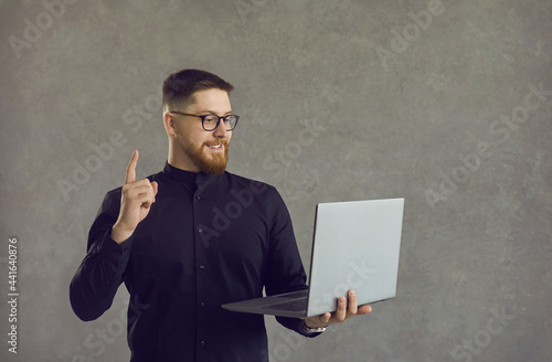 Happy intelligent young man in glasses standing against grey studio background holding laptop computer, looking at screen, smiling and pointing index finger up, struck by smart idea of online business