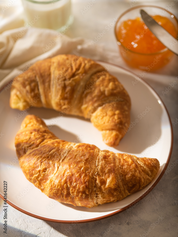 Breakfast table. There are two croissants with a beautiful brown crust on a white plate. In the background, in a glass vase, orange jam. Light background. High angle view.