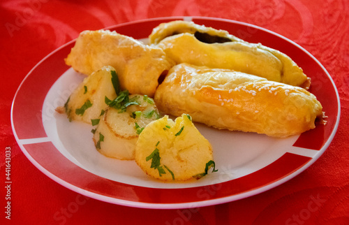 Plate served on table with red tablecloth, plate of baked rolls of spinach and ham served with potatoes seasoned with parsley.