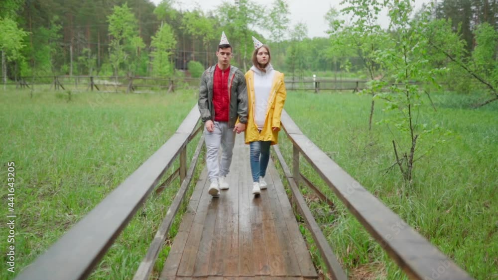 Sad unhappy young couple in raincoats and party hats walking down wooden path in countryside in rain in slow motion. Ruined dreams and broken expectations concept Sad unhappy young couple in raincoats and party hats walking down wooden path in countryside in rain in slow motion. Ruined dreams and broken expectations concept