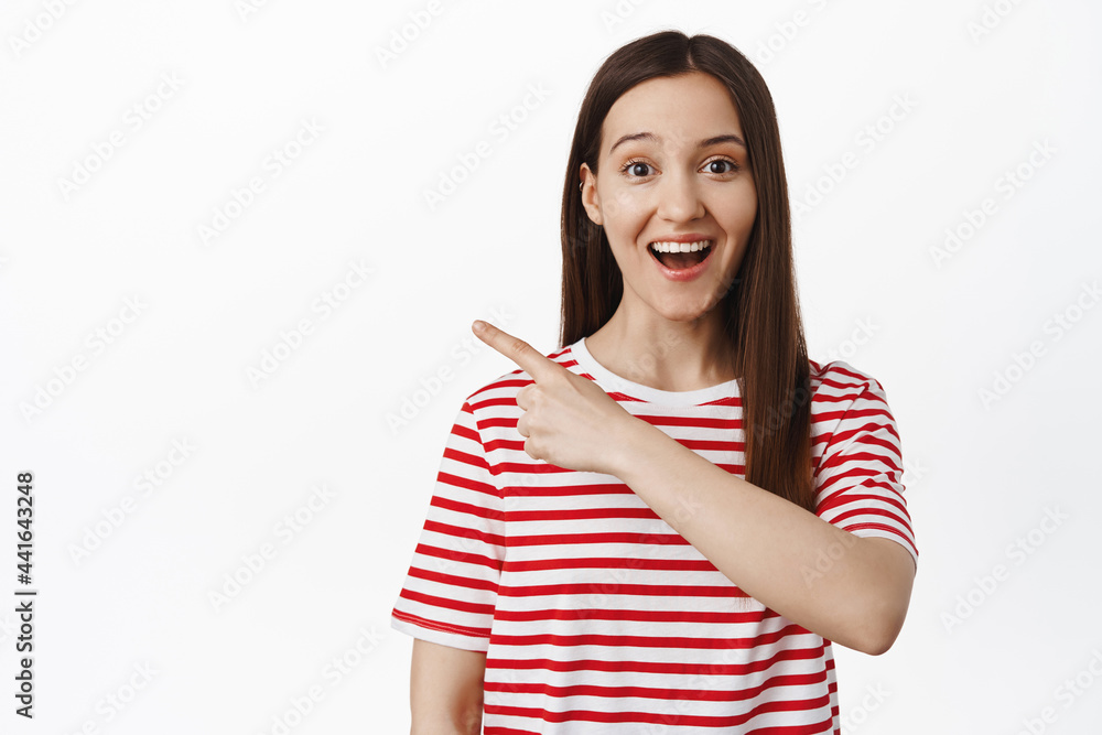 Portrait of young woman looks excited, smiling and pointing finger left at sale, showing advertisement logo, standing in red striped t-shirt against white background Portrait of young woman looks excited, smiling and pointing finger left at sale, showing advertisement logo, standing in red striped t-shirt against white background