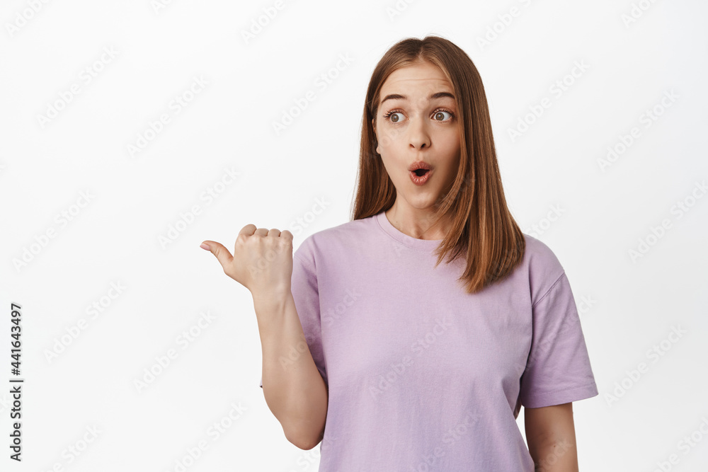Wow this is good. Natural blond girl pointing and looking left with impressed and thrilled face, checking out promo deal, event poster or logo on white empty space, studio shot Wow this is good. Natural blond girl pointing and looking left with impressed and thrilled face, checking out promo deal, event poster or logo on white empty space, studio shot