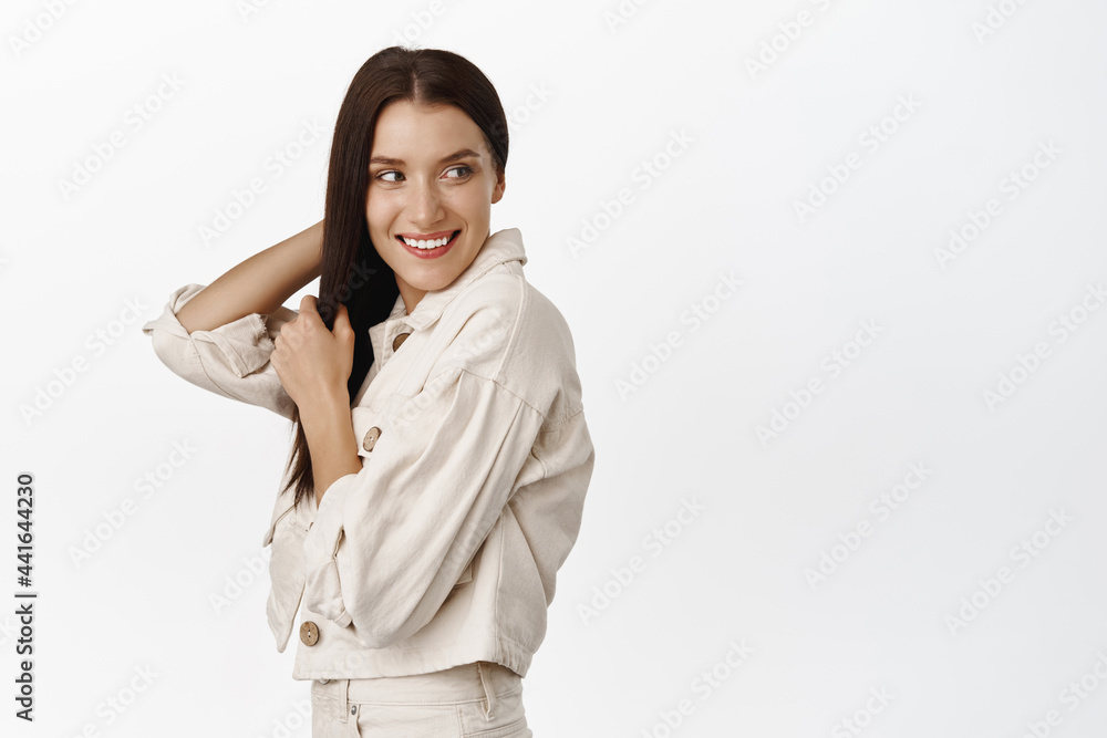 Beautiful adult woman turn back, looking behind her shoulder with carefree smile, reading promo text, touching dark hair, standing over white background Beautiful adult woman turn back, looking behind her shoulder with carefree smile, reading promo text, touching dark hair, standing over white background