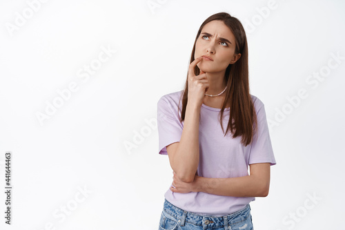 Hmm let me think. Young woman looks up thoughtful, touch lip and thinking, making choice, deciding what to order takeaway, standing against white background