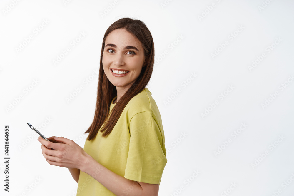 Portrait of young candid girl, smiling natural, using smartphone, holding cellphone and looking happy at camera, chatting, standing against white background Portrait of young candid girl, smiling natural, using smartphone, holding cellphone and looking happy at camera, chatting, standing against white background