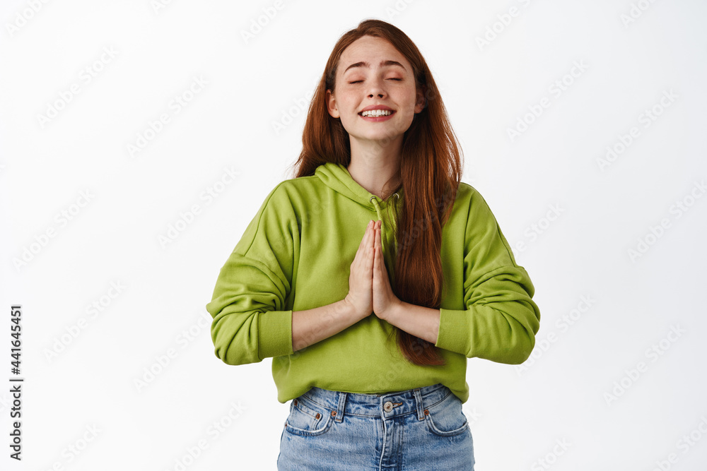 Hopeful redhead woman begging, praying with hands pressed together, making wish and smiling, anticipating miracle, standing over white background Hopeful redhead woman begging, praying with hands pressed together, making wish and smiling, anticipating miracle, standing over white background