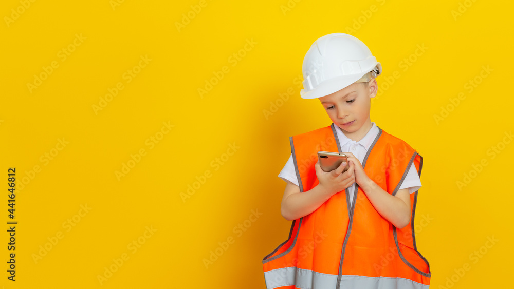 A child builder (engineer) dressed in a reflective vest and a construction helmet stands on a yellow background and makes a business call from his smartphone. A child builder (engineer) dressed in a reflective vest and a construction helmet stands on a yellow background and makes a business call from his smartphone.