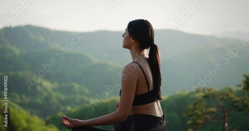 Slender young woman practicing yoga outdoors, meditating in mountains, sitting relaxed in lotus position with closed eyes.