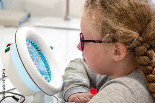 A girl is sitting in front of a device for stimulating vision.Ophthalmic equipment.The concept of vision treatment children.Orthoptic and Pleoptic Therapy.Concept of recovery and training of vision