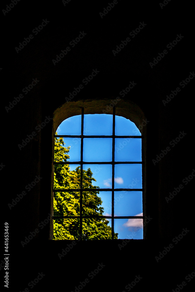The window is closed with a grill. On a dark background of the wall, an open window. Through the bars on the window, you can see the blue sky and green leaves of the tree.