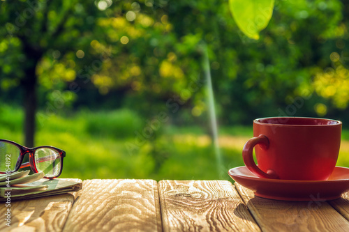 A cup of hot coffee and glasses with a newspaper on a wooden table in a green garden in the morning. Summer background