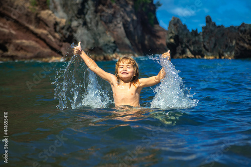 Amazed child playing and splashing in the sea. Kid having fun outdoors. Summer vacation and healthy family lifestyle concept. Little boy raises hands up in the water and splashes water drops.