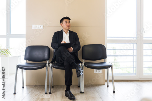 Young successful Asian man, waiting for a job interview, in the waiting room of the office center, sitting on a chair near the reception of the office center