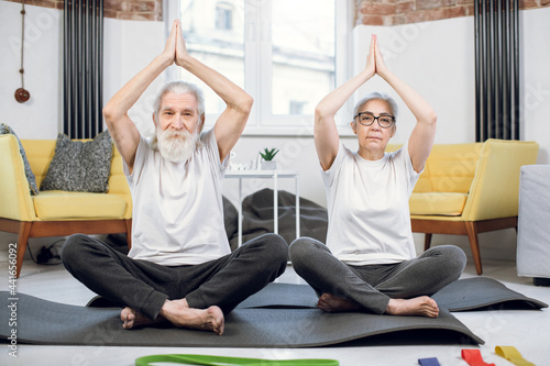 Obraz na plátně Happy aged man and woman practising yoga at home while sitting in lotus position with namaste hands above heads