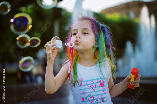 pretty little girl with colorful dyed hair blowing soap bubbles. Splashing city fountain is on background. Saint-Petersburg  Russia.Image with selective focus 