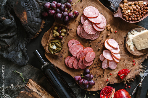 Antipasto cold meat platter with sausage, ham, salami, decorated with cheese, fruits, bottle of wine, nuts and olives on wooden board over black background. Meat appetizer, top view