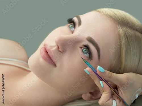 Eyelash Extension Procedure. Close up of beauty model's face during procedures of eye lash extension and master’s hand with tweezers, selected focus.