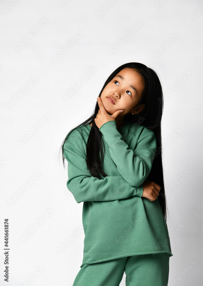 Thoughtful pensive asian preteen girl wearing green clothing, standing against studio background looking up and thinking on idea or problem making decision choosing solution touching chin by hand Thoughtful pensive asian preteen girl wearing green clothing, standing against studio background looking up and thinking on idea or problem making decision choosing solution touching chin by hand