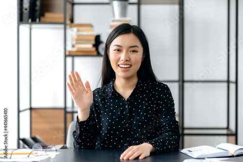 Webcam headshot of a positive pretty, smart confident young asian woman, freelancer or manager, sits at work desk, talking with colleagues or friends via video link, waving hand, smiling friendly