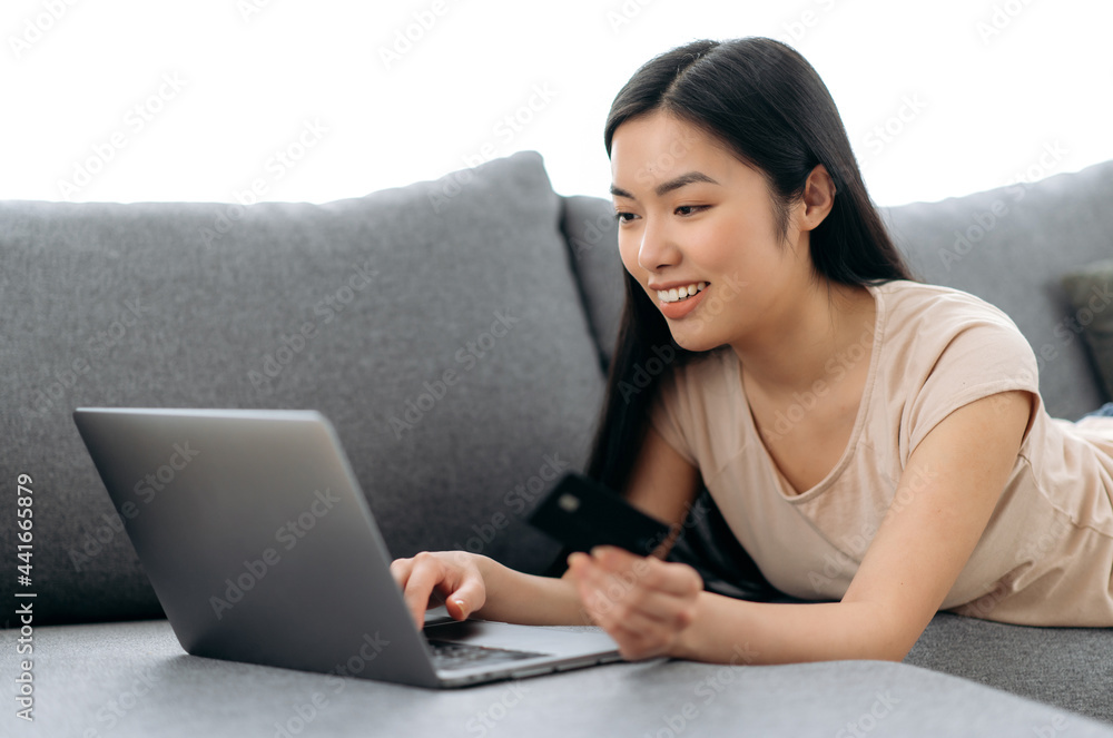 Online shopping. Positive beautiful young asian brunette woman lying on a couch, uses laptop and credit card to pay for online purchases and delivery, enters data, looks at the screen, smiles Online shopping. Positive beautiful young asian brunette woman lying on a couch, uses laptop and credit card to pay for online purchases and delivery, enters data, looks at the screen, smiles