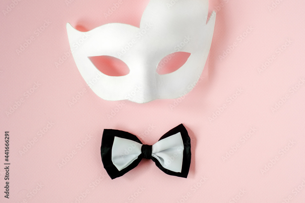 A black and white bow tie and a white carnival mask on a pastel pink background. Copy space.