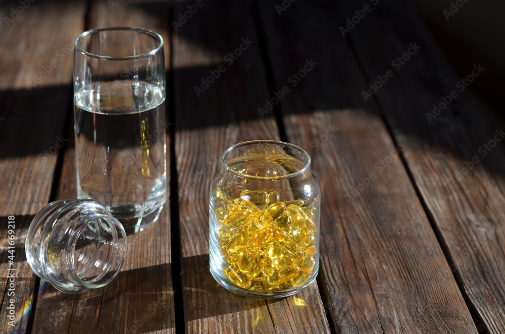 Glass jar with vitamins in yellow gelatin capsules and a glass of water. Glass jar with vitamins in yellow gelatin capsules and a glass of water.