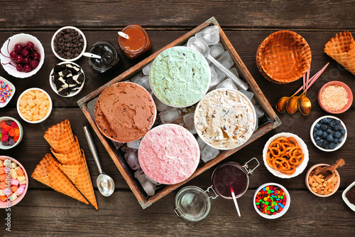 Summer ice cream buffet with a mixture of flavors and sweet toppings. Top view table scene on a dark wood background.